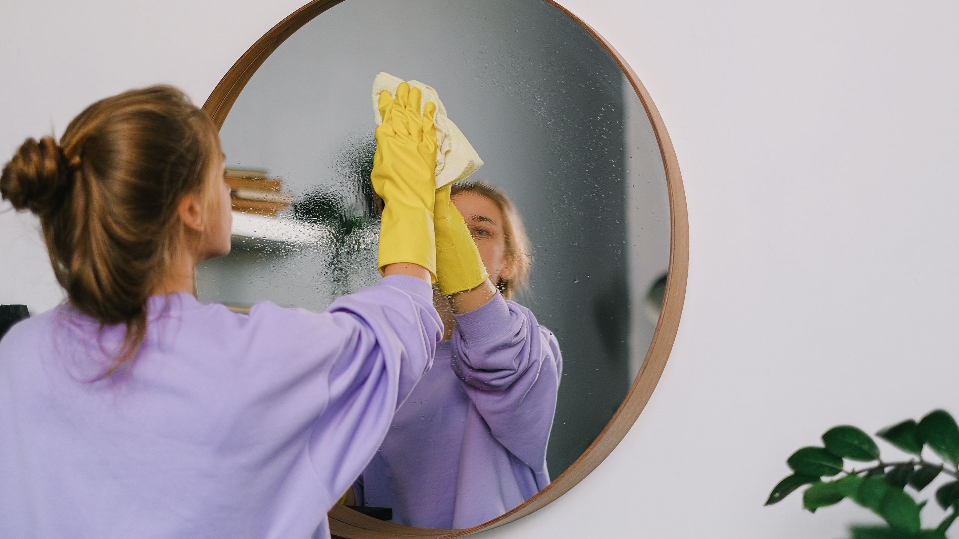 Person in purple shirt cleaning round mirror with yellow rubber gloves