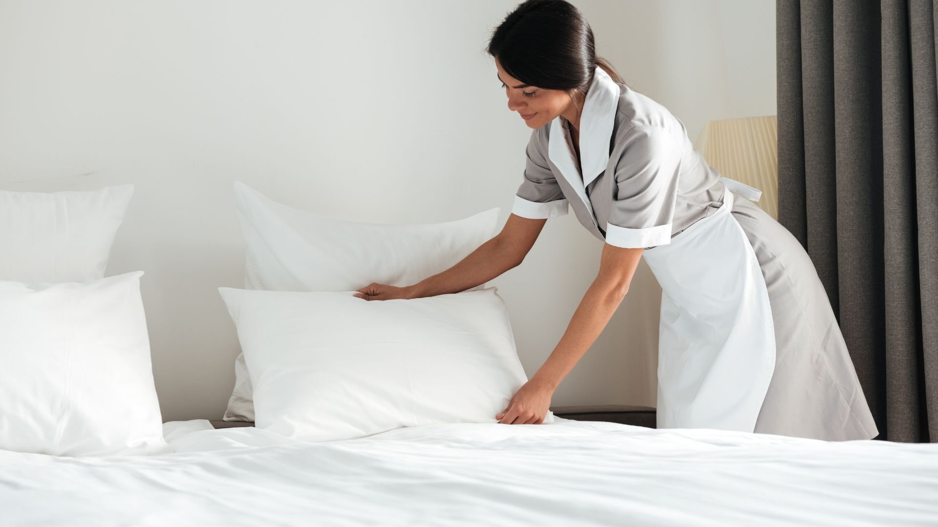 Housekeeper in uniform making a bed with white linens in a hotel room